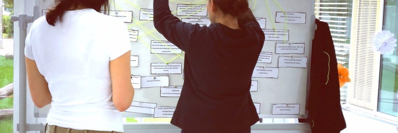 Das Bild zeigt zwei Personen von hinten, mutmaßlich Lehrerinnen des SFZ München West, die vor eine Moderationstafel stehen.