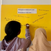 zwei Schülerinnen stehen vor einem gelben Plakat und eine der beiden schreibt ihre Vervollständigung zu den Satzanfängen "Philosophieren bedeutet bzw. ist für mich..." auf dieses Plakat